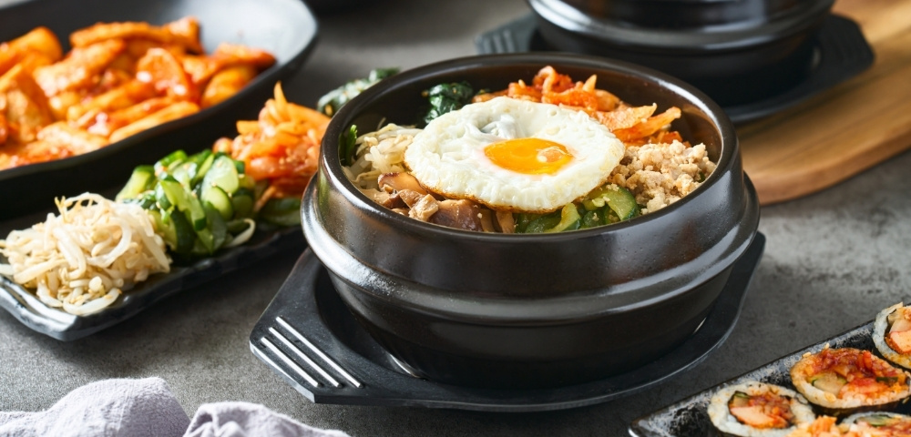 Steaming Korean bibimbap in a black stone bowl, topped with fried egg and vegetables, surrounded by colorful banchan and sushi rolls.