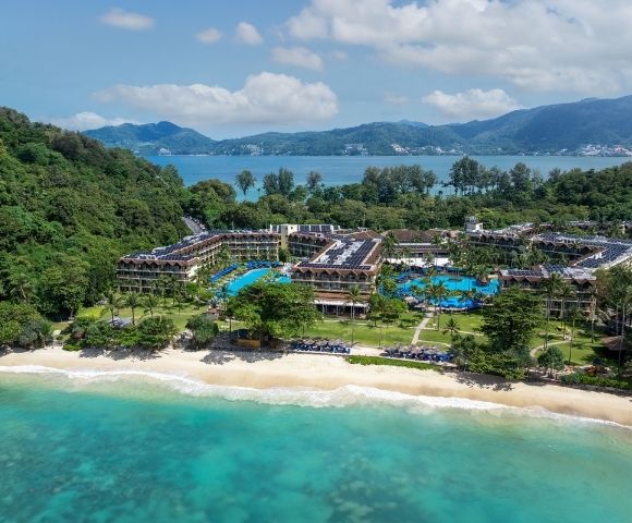 Aerial view of a sprawling beach resort complex with multiple buildings, swimming pools, and lush green grounds, situated between a sandy beach and a dense, tree-covered hillside, overlooking a calm turquoise ocean and distant mountains under a partly cloudy sky.