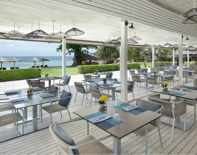 An outdoor dining area with many tables and chairs set under a white covered patio with a view of the ocean and beach.