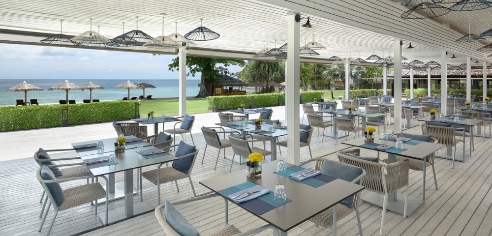 An outdoor dining area with many tables and chairs set under a white covered patio with a view of the ocean and beach.