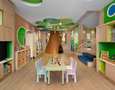 A colorful and inviting children's playroom featuring a large, wooden tree structure in the center with a playhouse opening, a green leafy canopy overhead, a table with chairs, and shelves filled with toys and books.