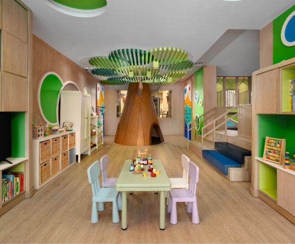 A colorful and inviting children's playroom featuring a large, wooden tree structure in the center with a playhouse opening, a green leafy canopy overhead, a table with chairs, and shelves filled with toys and books.