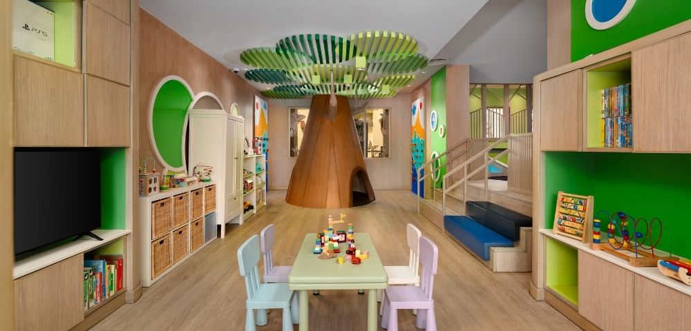 A colorful and inviting children's playroom featuring a large, wooden tree structure in the center with a playhouse opening, a green leafy canopy overhead, a table with chairs, and shelves filled with toys and books.
