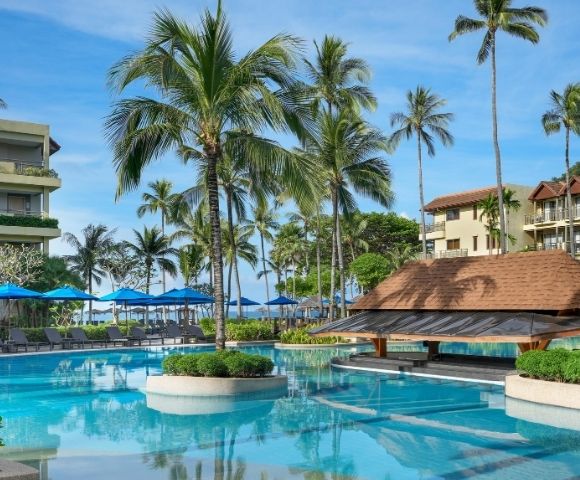 A tropical resort swimming pool with palm trees, lounge chairs, umbrellas, and a swim-up bar under a thatched roof, with buildings in the background.