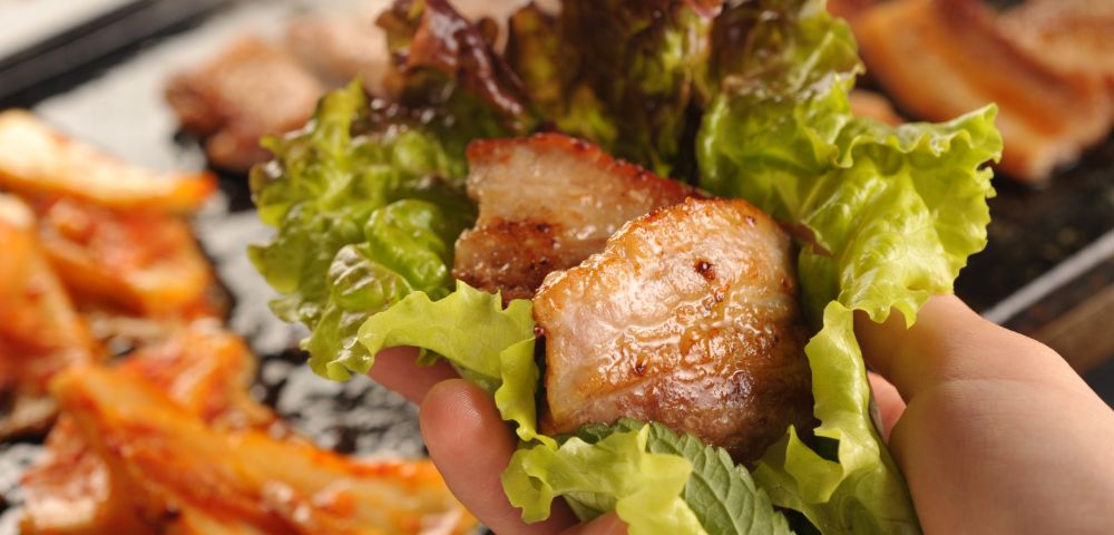 Hand holds a lettuce wrap filled with grilled pork belly, with kimchi and Korean barbecue dishes blurred in the background, warm and appetizing.