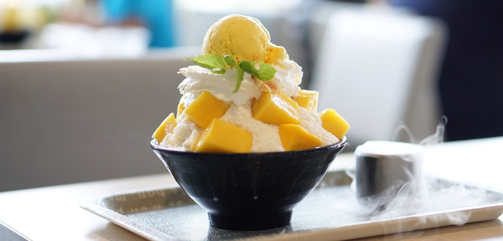 Bowl of mango shaved ice topped with whipped cream, mint, and vanilla ice cream, served on a café table with a warm, inviting tone.