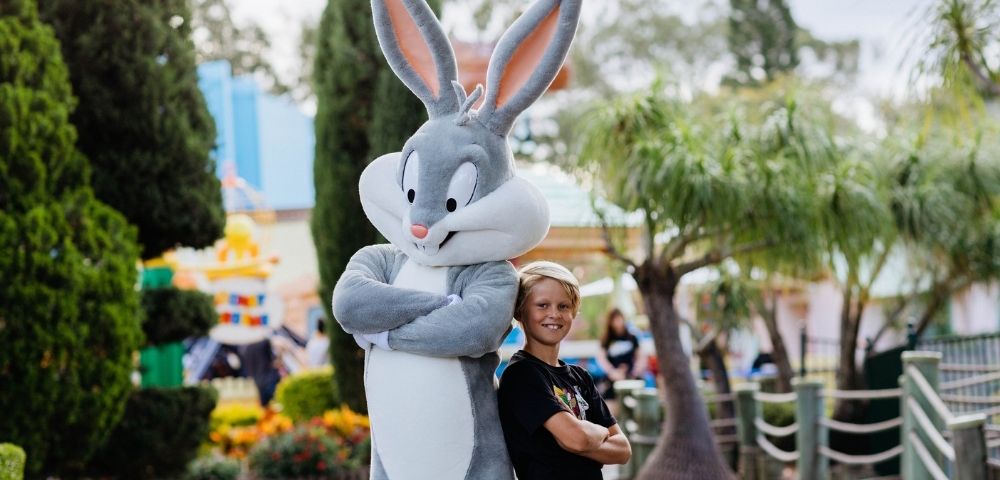 A child poses for a photo with a costumed Bugs Bunny character in an outdoor amusement park setting.