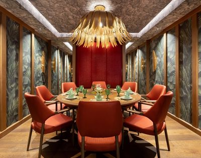 Round dining table set for a meal with coral chairs, set in a room with tropical leaf wallpaper and a dramatic floral chandelier.