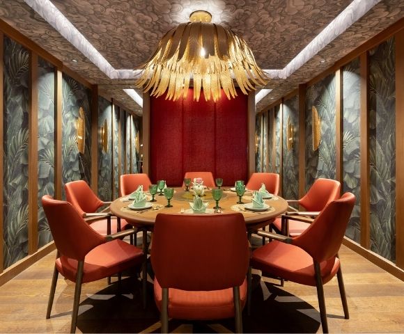 Round dining table set for a meal with coral chairs, set in a room with tropical leaf wallpaper and a dramatic floral chandelier.