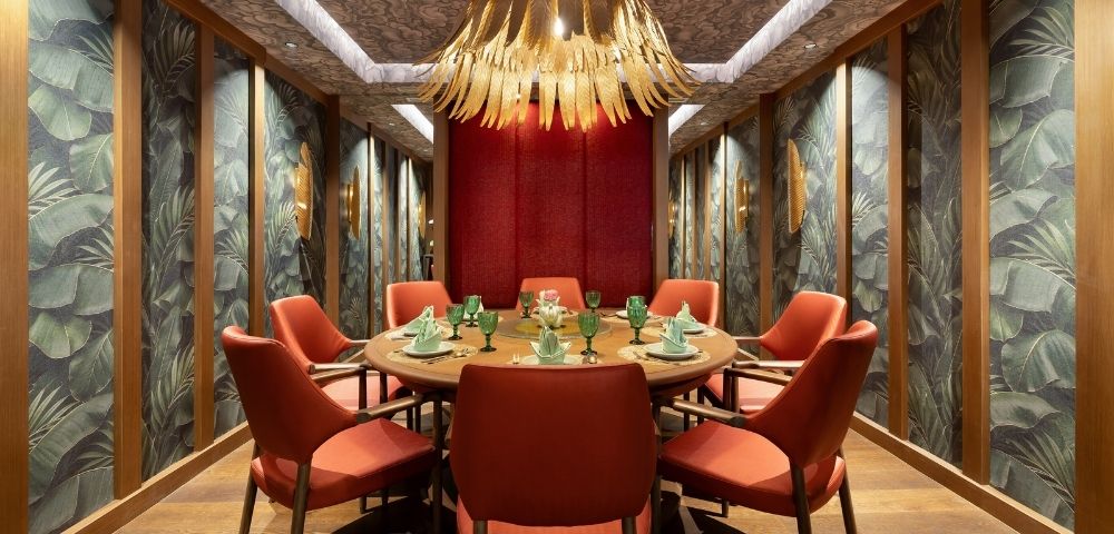 Round dining table set for a meal with coral chairs, set in a room with tropical leaf wallpaper and a dramatic floral chandelier.