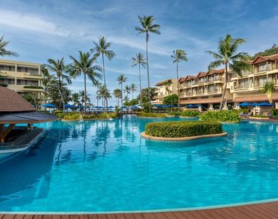 A large, resort swimming pool with bright blue water, surrounded by palm trees and multi-story hotel buildings with balconies.