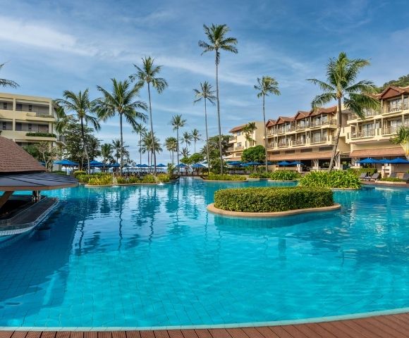 A large, resort swimming pool with bright blue water, surrounded by palm trees and multi-story hotel buildings with balconies.