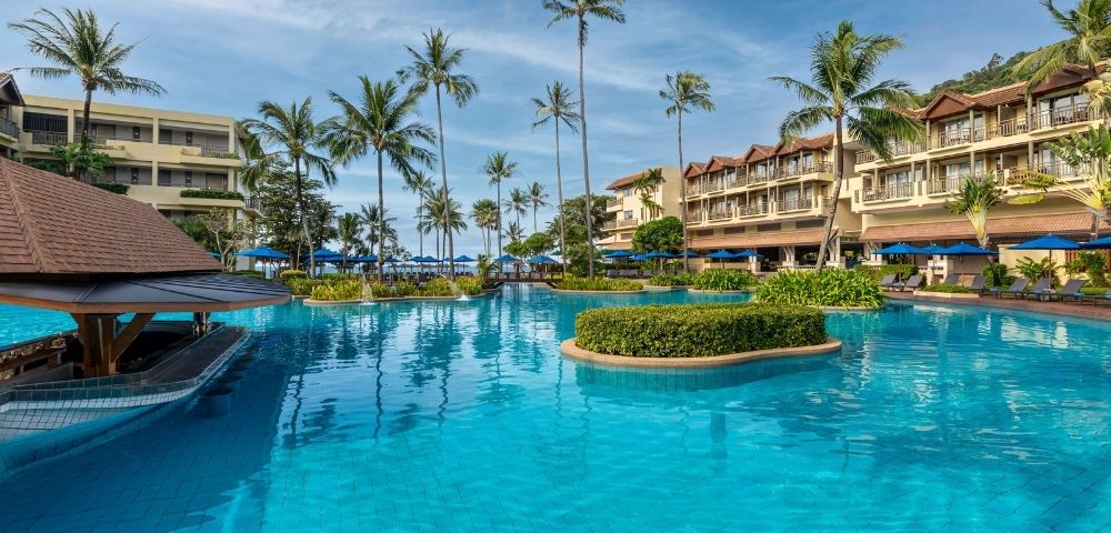 A large, resort swimming pool with bright blue water, surrounded by palm trees and multi-story hotel buildings with balconies.