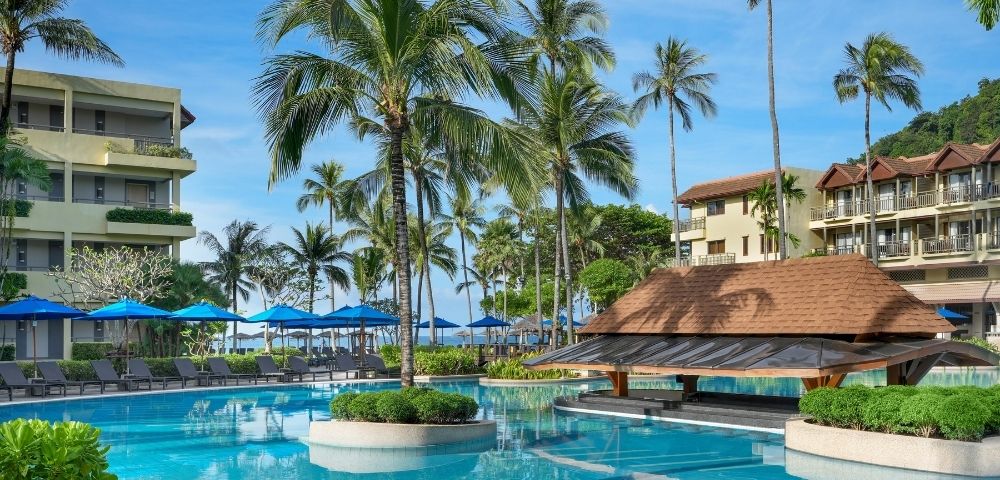 A resort swimming pool with lush palm trees and beachfront hotels under a clear blue sky.