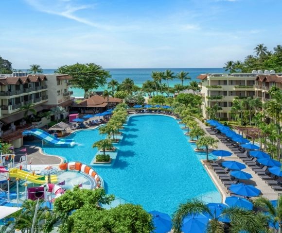 Aerial view of a tropical resort with a large blue swimming pool complex featuring waterslides and a splash pad, surrounded by lush palm trees, lounge chairs with blue umbrellas, and multi-story hotel buildings, with the ocean visible in the background.