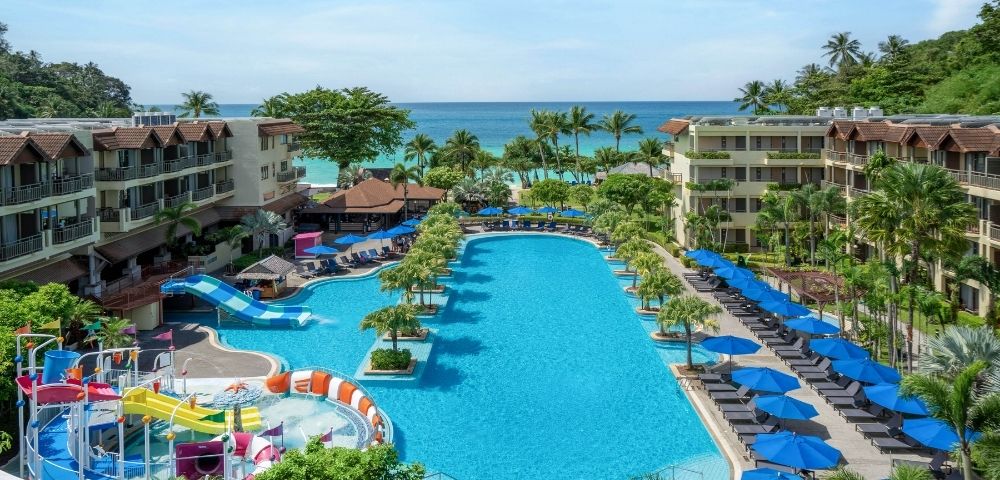 Aerial view of a tropical resort with a large blue swimming pool complex featuring waterslides and a splash pad, surrounded by lush palm trees, lounge chairs with blue umbrellas, and multi-story hotel buildings, with the ocean visible in the background.