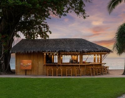 Thatched tropical beach bar with wooden stools and hanging lights, ocean in background at sunset.