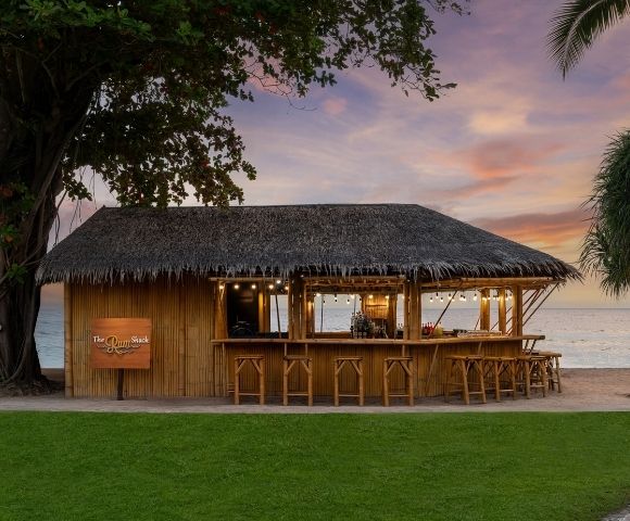 Thatched tropical beach bar with wooden stools and hanging lights, ocean in background at sunset.