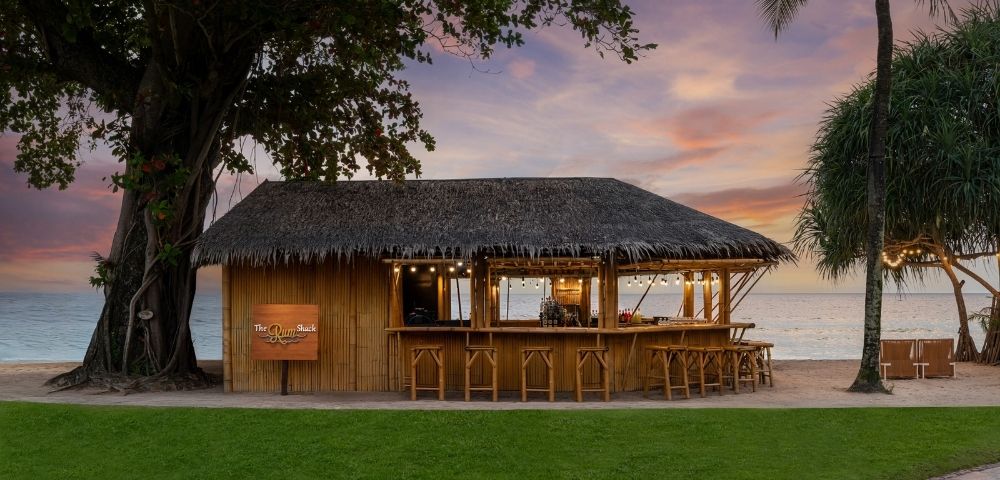 Thatched tropical beach bar with wooden stools and hanging lights, ocean in background at sunset.