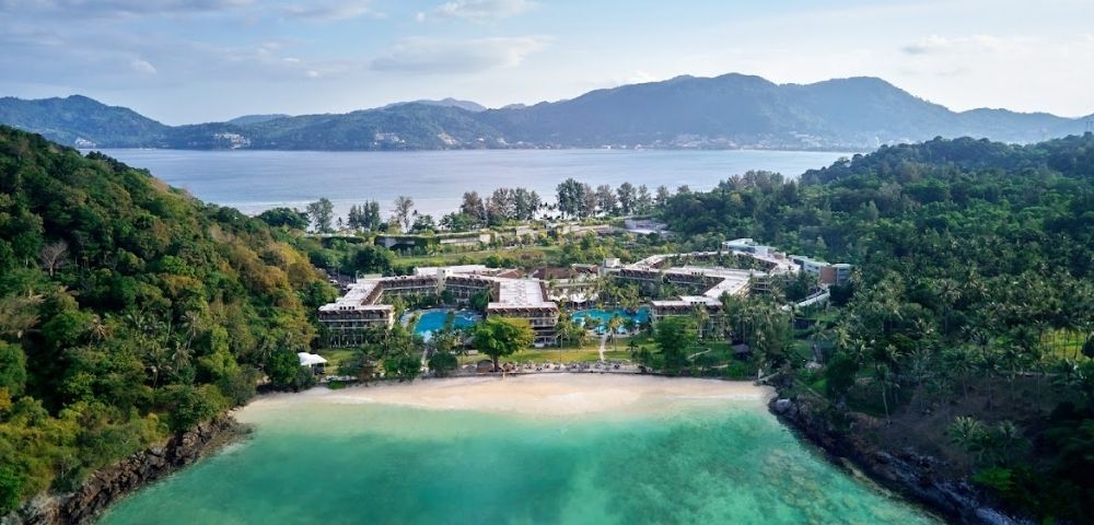 Aerial view of a beachfront resort surrounded by lush greenery, clear turquoise water, and mountains in the background under a partly cloudy sky.