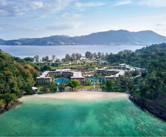 Aerial view of a beachfront resort surrounded by lush greenery, clear turquoise water, and mountains in the background under a partly cloudy sky.