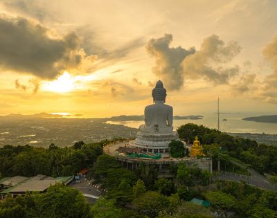 A colossal white Buddha statue sits atop a lush green hill overlooking a coastal city and the ocean during a vibrant sunset.