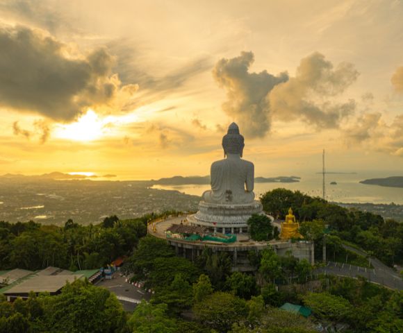 A colossal white Buddha statue sits atop a lush green hill overlooking a coastal city and the ocean during a vibrant sunset.