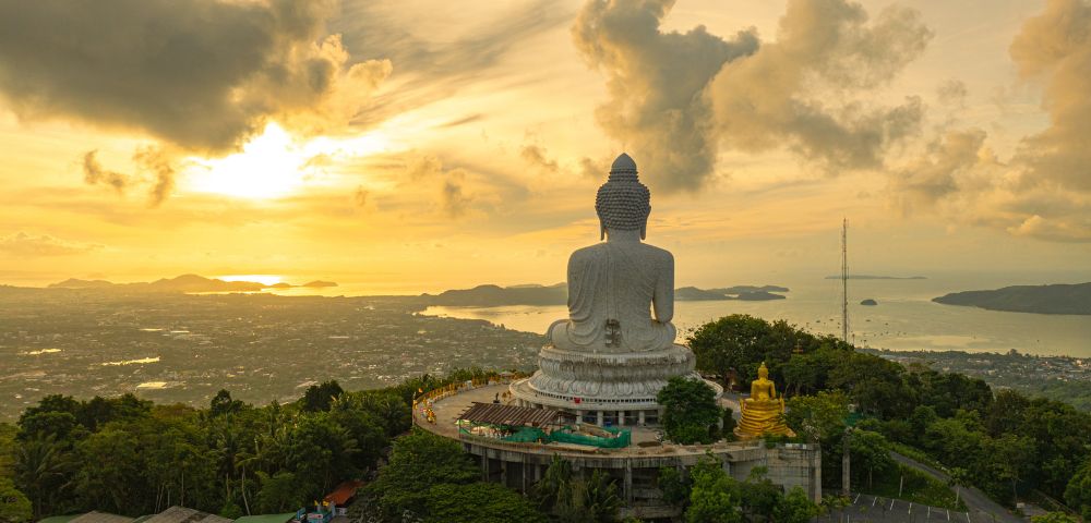 A colossal white Buddha statue sits atop a lush green hill overlooking a coastal city and the ocean during a vibrant sunset.