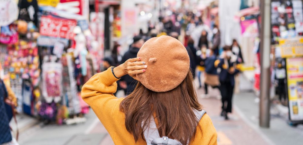 Woman seen from behind, walking through a busy, colorful shopping street in an upbeat city scene.