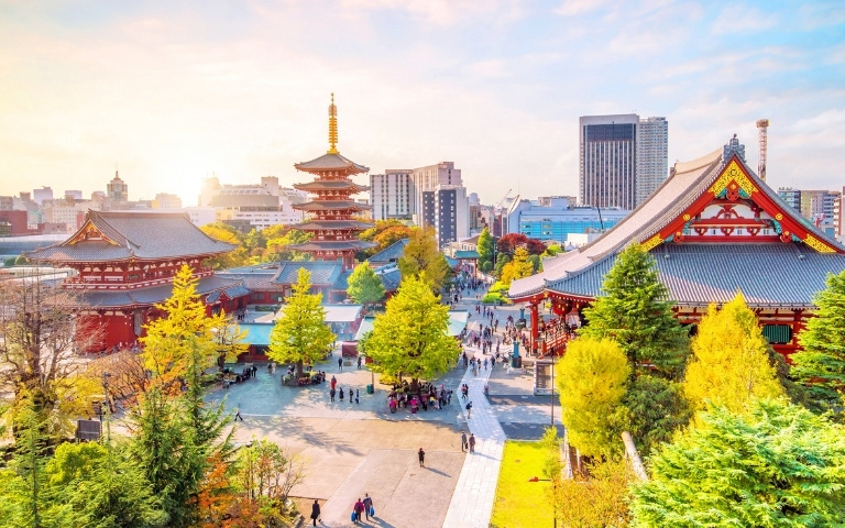 Panoramic view of Tokyo’s Asakusa district, with Senso-ji Temple, red pagoda, autumn trees, visitors, and modern skyline in warm sunlight.