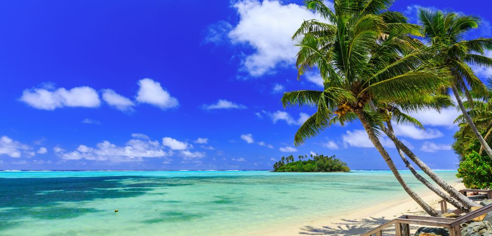 Tropical beach with clear turquoise water, white sand, leaning palm trees, and a distant small island under a vibrant blue sky with white clouds.