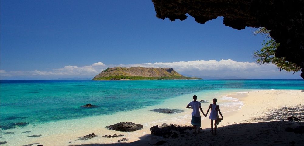 A couple walks hand-in-hand on a pristine white sand beach in front of a brilliant turquoise ocean, with a green, mountainous island in the distance.