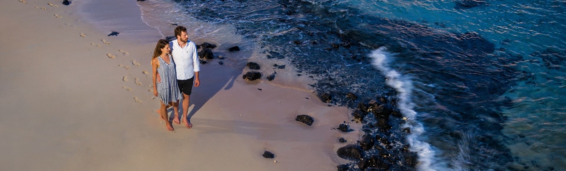 A couple strolls hand-in-hand along a sandy beach at the edge of the ocean.