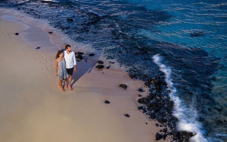 A couple strolls hand-in-hand along a sandy beach at the edge of the ocean.