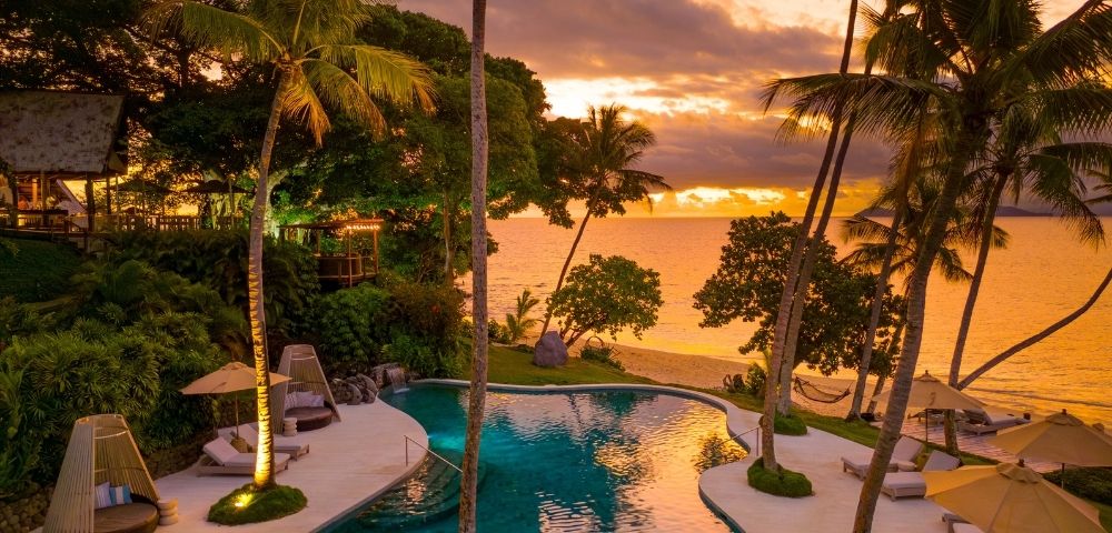 A serene tropical resort at sunset features a curved swimming pool overlooking a calm ocean, with palm trees, lounge chairs, umbrellas, and a distant hammock visible along the beach.