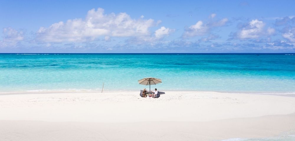 A couple enjoys a picnic lunch on a white sand beach under a large umbrella with the turquoise ocean and blue sky in the background.