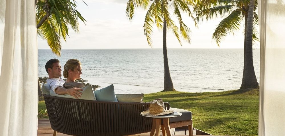 A couple sits on a wicker sofa, looking out at the ocean from a tropical beachfront resort.