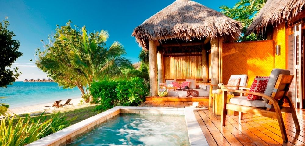 A tropical resort scene with a thatched-roof cabana, lounge chairs, and a private plunge pool overlooking a turquoise ocean and beach with overwater bungalows in the distance.