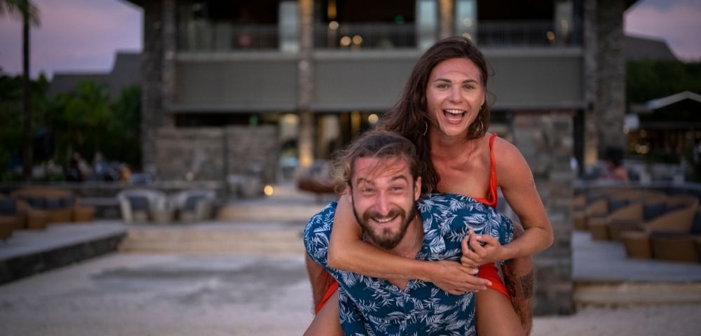 A man gives a woman a piggyback ride on a sandy beach in front of a blurred resort building.
