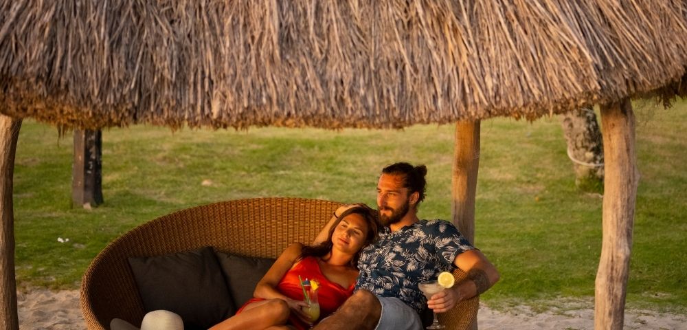 A couple relaxes on a round, rattan daybed under a thatched roof umbrella, holding tropical drinks with lemon and mint garnishes on a sandy beach with a grassy lawn in the background.