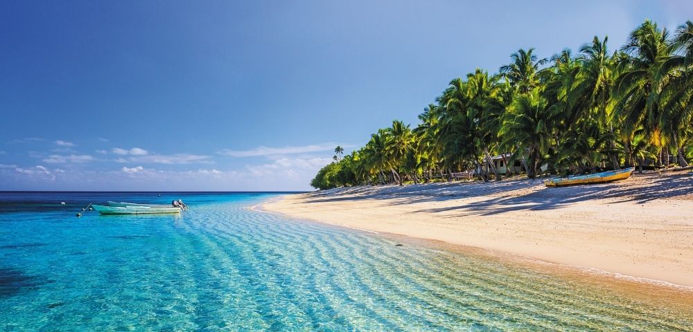 A pristine tropical beach with crystal-clear turquoise water, lined with lush green palm trees and a golden sand shore. Two small boats are visible in the shallow water, and another colorful boat rests on the sand.