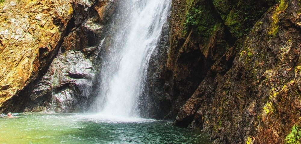 A powerful waterfall cascades down mossy rocks into a clear, tranquil pool where a person is swimming.