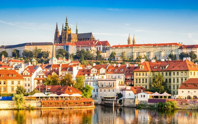 Sunny panoramic view of Prague across the Vltava River, with red-roofed historic buildings, Prague Castle, and St. Vitus Cathedral.
