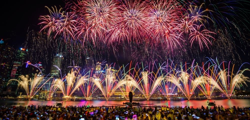 Spectacular fireworks and colorful fountains light up a waterfront skyline at night as a large crowd watches in celebration.