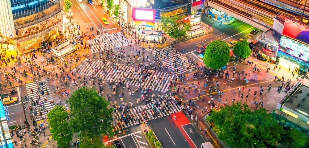 Aerial view of Tokyo’s Shibuya Crossing packed with pedestrians, traffic, glowing billboards, and tree-lined streets in a vibrant city scene.