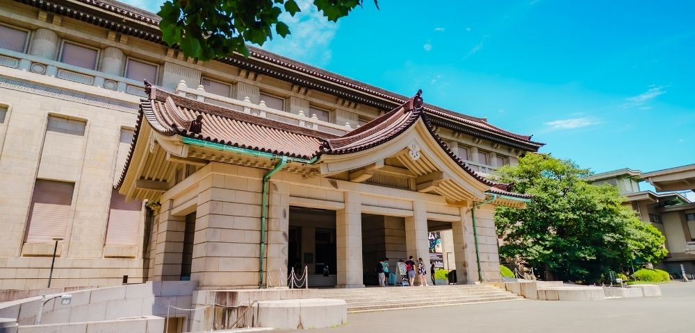 Grand historic museum entrance with ornate Japanese-style tiled roof, stone columns, and visitors beneath a bright blue sky; serene, welcoming scene.