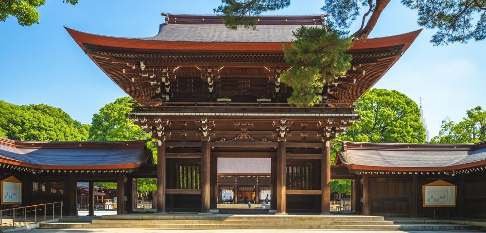 Grand wooden Japanese temple gate with layered tiled roofs, ornate carvings, and open side wings, framed by green trees under blue sky.
