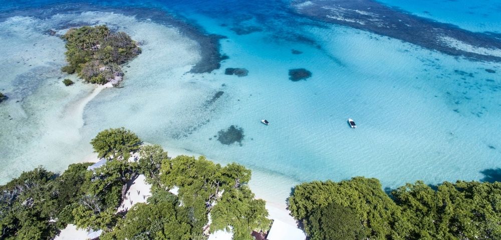 Aerial view of a tropical coastline with lush green trees, white sand beaches, and crystal-clear turquoise water. Two small boats are visible in the calm sea.