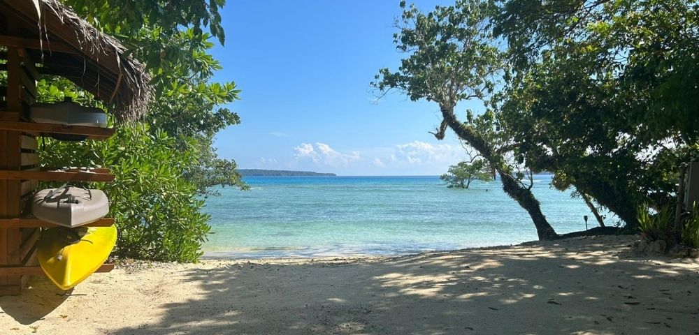 A thatched-roof shelter frames a tropical beach scene with a yellow kayak and two gray kayaks stored on wooden shelves. The turquoise water is calm and clear, leading to a distant island under a bright blue sky with fluffy clouds. Lush green trees overhang the sandy shore, casting dappled shadows.