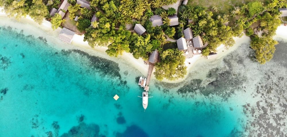 Aerial view of a tropical island resort with bungalows nestled among lush green trees, a white sandy beach, a wooden pier with boats, and crystal-clear turquoise waters revealing coral reefs below.